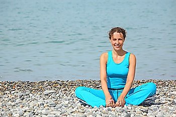 smiling woman is stretching on a coast of sea. she is wearing sporty clothers.