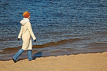 Girl walks on autumn beach in the hat, coat, jeans and sports shoes.