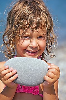 smiling pretty little girl holds in hands pebble on seacoast in afternoon