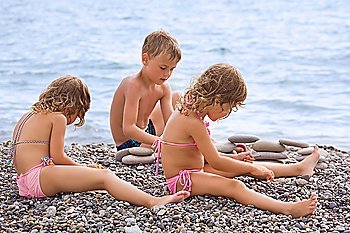 children three together sitting on stony beach, creates pyramid from pebble