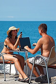 Man and woman sitting at table under open sky on background of sea. Man works for laptop. Vertical format.