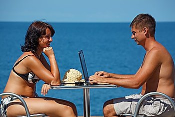 Man and woman sitting at table under open sky on background of sea. Man works for laptop.