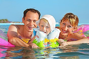 Happy family with little girl in white hat and lifejacket bathing in pool against sea