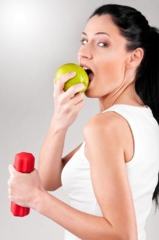 sporty woman is biting off green apple on grey background