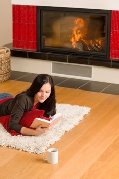 Happy young woman lying by fireplace on carpet reading book