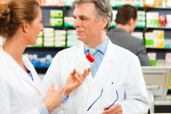 Two pharmacists with pharmaceuticals in hand consulting each other in a pharmacy; a client is standing in the background