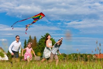 Happy family - mother, father, children - running over a green meadow in summer; they are flying a kite