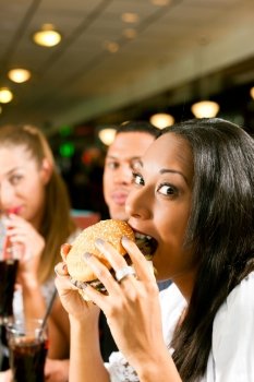 Friends - one couple is African American - eating hamburger and drinking soda in a fast food diner; focus on the woman in front