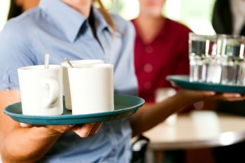 Woman as waitress in a bar or restaurant with coffee mugs; in the background are guests