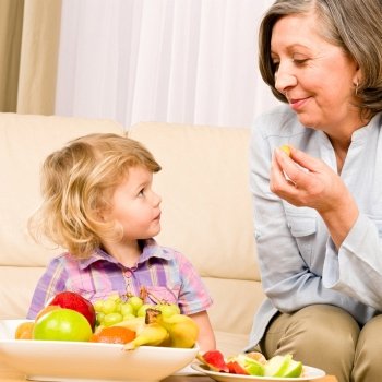 Little girl watch grandmother eat fruit relaxing on sofa