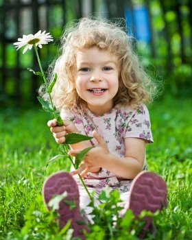 Happy smiling child with flower sitting on green grass outdoors in spring garden