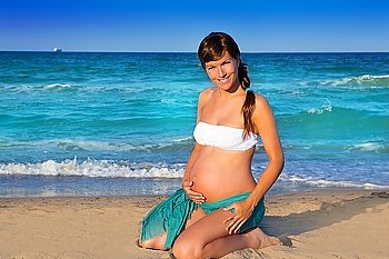 Beautiful pregnant woman on knees at blue beach sand in summer vacation