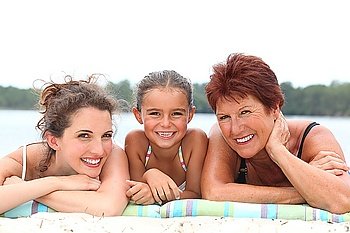 a 30 years old woman, a little girl and a 55 years old woman lying down on the beach, behind sea and forest background