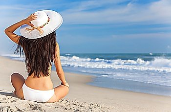 A sexy young brunette woman or girl wearing a white bikini and sun hat sitting on a deserted tropical beach with a blue sky