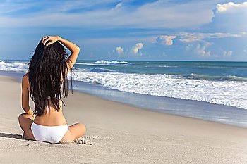 A sexy young brunette woman or girl wearing a white bikini sitting on a deserted tropical beach with a blue sky