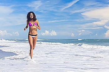 A sexy young brunette woman or girl wearing a bikini running through the surf on a deserted tropical beach with a blue sky