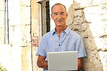 Senior man with laptop in front of stone house