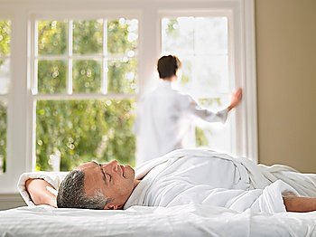 Man relaxing in bed wife looking through window in background