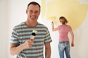 Smiling man holding paintbrush while woman paints wall with roller