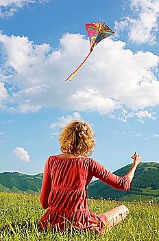 Woman sitting in mountain field Flying Kite, back view