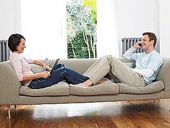Couple reclining on couch using laptop and cell phone