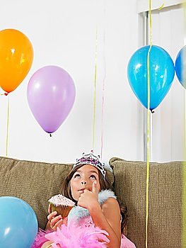 Young girl (7-9) sitting on sofa eating cupcake looking up at balloons