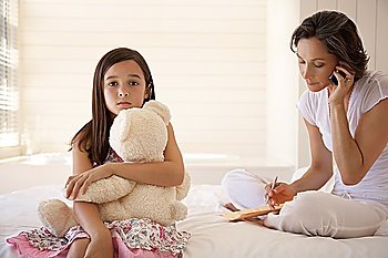 Mother using cell phone daughter cuddling teddy sitting on bed