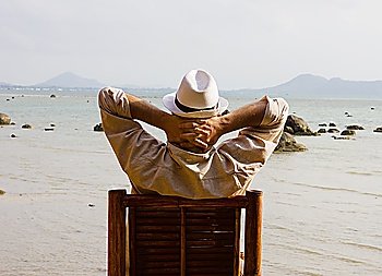 man sitting on a chair and looks at the sea holding his hands on his neck, back view