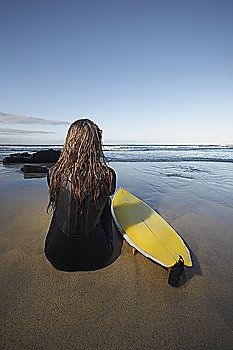 Woman sitting by surfboard on beach, back view
