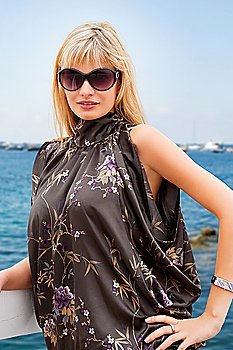 young blond beautiful woman in elegant dress and sunglasses near the sea looking in camera