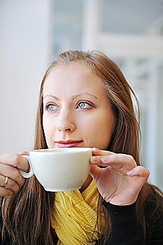 young woman holding  cup of coffee and looking out  window. She drinks coffee at  cafe