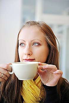 young woman holding  cup of coffee and looking out  window. She drinks coffee at  cafe