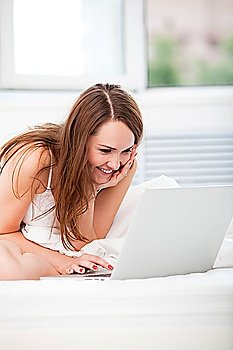 Beautiful happy young woman sittting in bed and using laptop at home