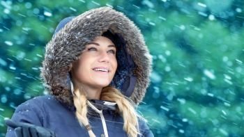 Portrait of a nice woman in winter park enjoying falling snow, wearing warm coat with fur hood, happy wintertime holidays 