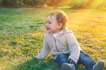 Cute baby boy outdoors, sweet little child sitting on fresh green grass field in bright sunny day, enjoying first spring days, happy carefree childhood. Cute baby boy outdoors