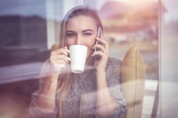 Portrait of a pretty blond woman with pleasure talking on the phone and drinking coffee, looking through the window and enjoying nature view. Calm female on the phone