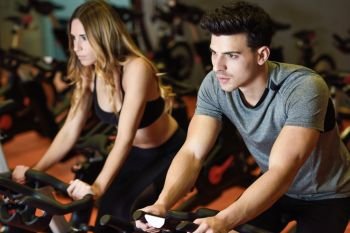 Young man and woman biking in the gym, exercising legs doing cardio workout cycling bikes. Two people in a spinning class wearing sportswear.