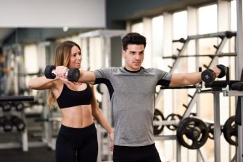 Female personal trainer helping a young man lift dumbells while working out in a gym