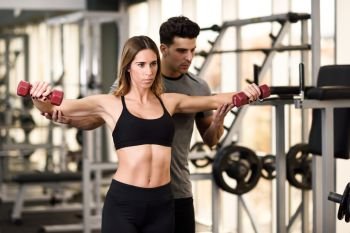Personal trainer helping a young woman lift dumbells while working out in a gym