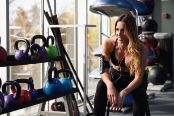 Young woman listening to music with smartphone sitting at gym. Young fitness female using headphones with dumbbells at the background.