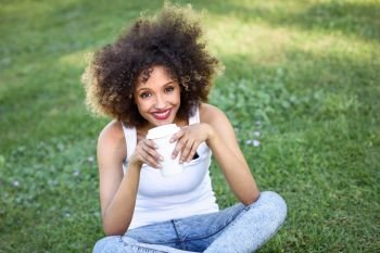 Beautiful young African American woman with afro hairstyle. Girl drinking coffee in park sitting on grass wearing casual clothes smiling.