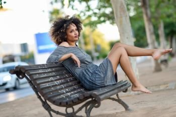 Young black woman with afro hairstyle sitting on a bench in urban background moving her legs. Mixed girl wearing casual clothes