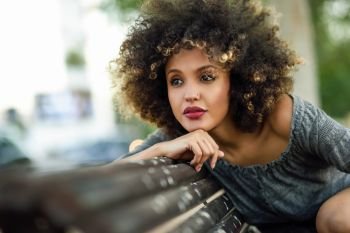 Young black woman with afro hairstyle sitting on a bench in urban background. Mixed girl wearing casual clothes