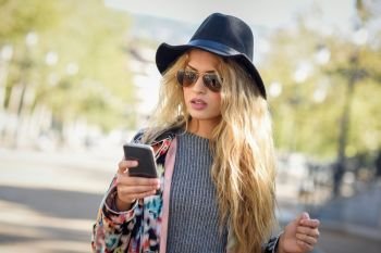 Young woman looking at her smartphone in urban background. Girl wearing jacket, hat, sweater and sunglasses.
