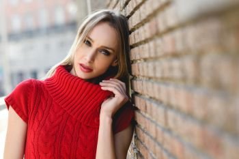 Close-up portrait of young blonde woman standing in the street near a brick wall. Beautiful girl in urban background wearing red dress. Female with straight hair and blue eyes.