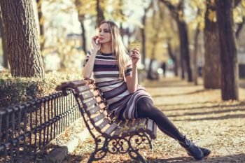 Young blonde woman sitting on a bench in the street of a park with autumn colors. Beautiful girl in urban background wearing striped dress and black tights. Female with straight hair holding an autumn leaf.