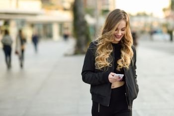 Blonde woman texting with her smart phone in urban background. Beautiful young girl wearing black jacket walking in the street. Pretty russian female with long wavy hair hairstyle.