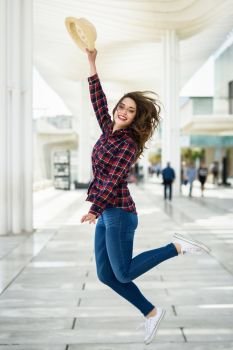 Young woman with beautiful blue eyes wearing plaid shirt and sun hat. Girl jumping in urban background.