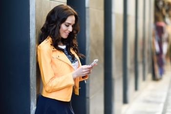 Young brunette woman, model of fashion, wearing orange modern jacket and blue skirt, texting with her smartphone in urban background.