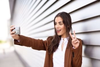 Young woman taking selfie photograph with smartphone in urban background. Beautiful girl wearing formal wear using smart phone. Young female with brown jacket smiling in urban background.
